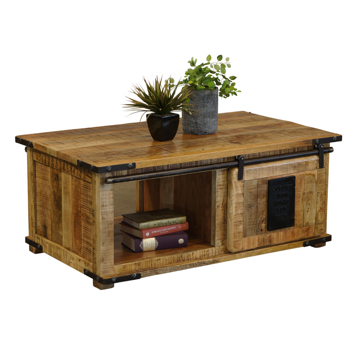 A rustic reclaimed wood coffee table with black metal hardware, featuring an open shelf with books and a sliding door with a decorative plaque. Two potted plants sit on top, set against a plain white background.