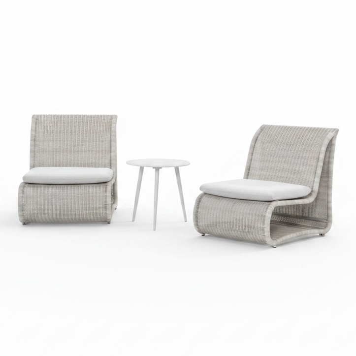 Two modern outdoor lounge chairs with light gray woven frames and white cushions, positioned on a poolside patio with a small round side table between them. A stone wall and green trees are visible in the background.