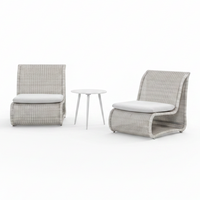 Two modern outdoor lounge chairs with light gray woven frames and white cushions, positioned on a poolside patio with a small round side table between them. A stone wall and green trees are visible in the background.