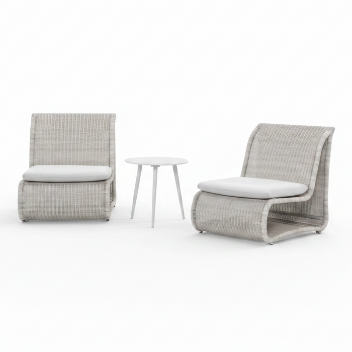 Two modern outdoor lounge chairs with light gray woven frames and white cushions, positioned on a poolside patio with a small round side table between them. A stone wall and green trees are visible in the background.
