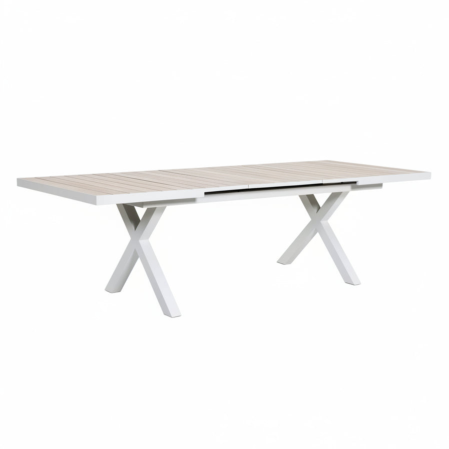 A rectangular outdoor dining table with a light beige slatted top and white X-shaped legs, shown on a tiled patio in a landscaped garden setting.