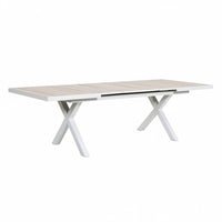 A rectangular outdoor dining table with a light beige slatted top and white X-shaped legs, shown on a tiled patio in a landscaped garden setting.