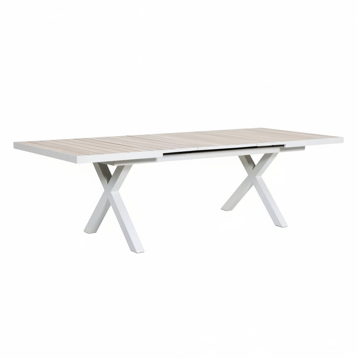 A rectangular outdoor dining table with a light beige slatted top and white X-shaped legs, shown on a tiled patio in a landscaped garden setting.