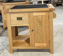 A small wooden kitchen bench cabinet or side table made of natural oak wood, featuring a single drawer with a brass pull, a lower open shelf, and a dark rectangular top surface.